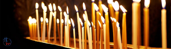 Row of tall lit candles in a holder, glowing warmly against a dark background in a solemn setting.