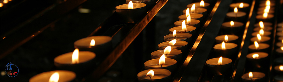 Rows of small lit candles in metal holders glowing warmly in a dark, contemplative setting.