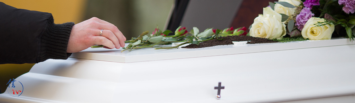 Close-up of a hand placing a flower on a white coffin adorned with roses, greenery, and a cross.